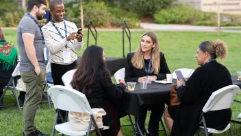Three attendees socializing at a table with a black cloth. Two attendees standing and smiling, showing something on their phones.