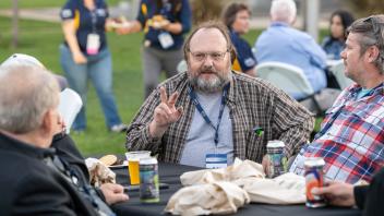 Attendees sitting at a table, conversing and smiling. One attendee raises their hand in a peace sign.