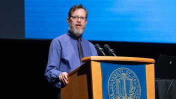Andy Jones MC'ing at a podium with a UC Tech 2024 background.