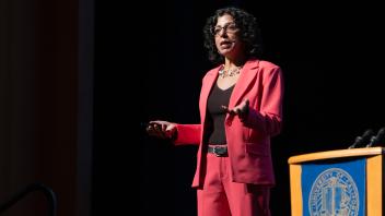 Keynote speaker, Raissa D'Souza, giving her keynote address at UC Tech. She is wearing a red suit with her arms raised in front of her.