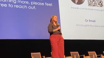 Kate McLoughlin giving her spotlight talk. She is standing with her presentation slides projected in the background.